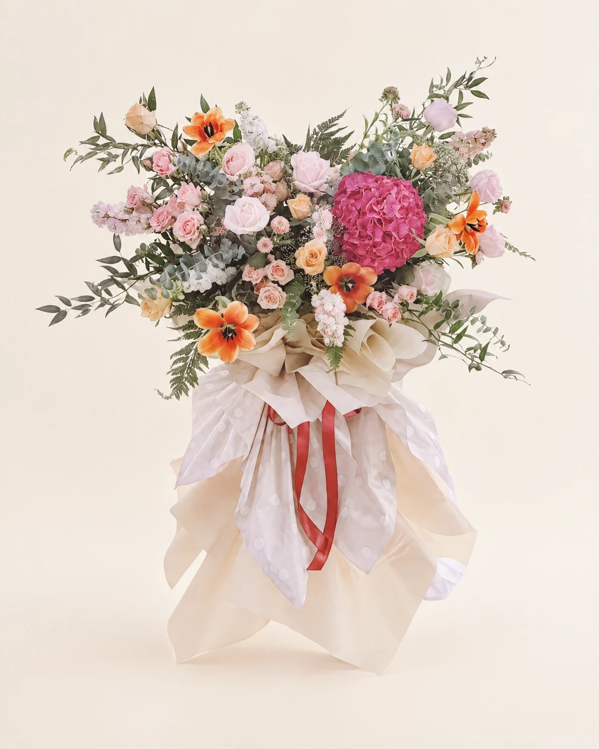 混合花卉花束(象牙色包裝) — 粉紅及桃色玫瑰、粉紅繡球花、橙色花材、白色及淡粉色填充花材、桉樹及其他綠葉、米白色包裝紙及紅色絲帶